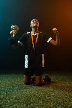 Adult female athlete kneeling on green turf holding gold cup in black jersey shorts orange medal