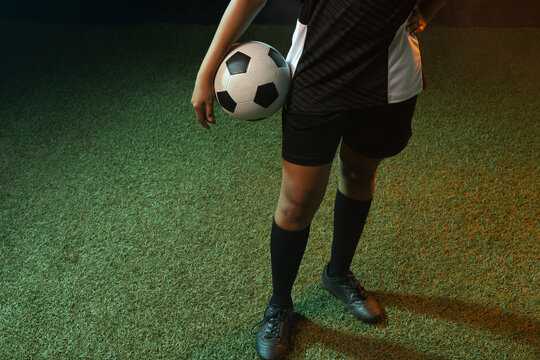 African American woman standing on turf holding black-white soccer ball at hip, in kit and cleats