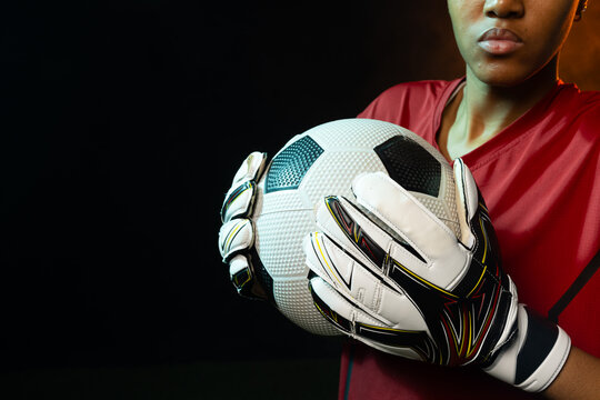 African American woman holding white ball with black panels wearing red jersey and gloves in studio