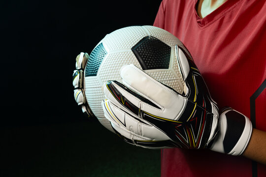 African American female goalkeeper in red kit holding white black soccer ball with gloves near turf