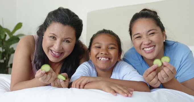 Female family reclining with child on white bed starting holding cucumbers pressing eyes laughing
