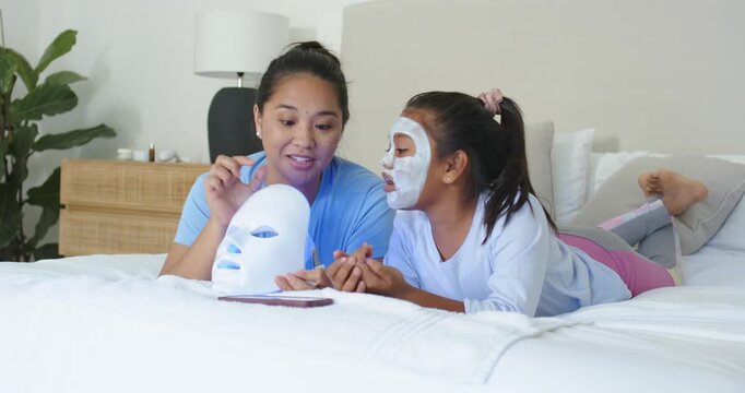 Diverse mother toggling LED mask, guiding child wearing sheet mask on bed for skincare via phone