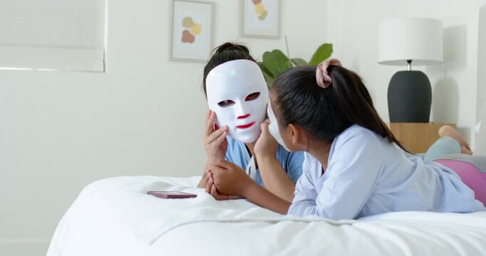 Diverse mom and daughter on bed, mom lifting LED mask, daughter touching, testing mask with phone