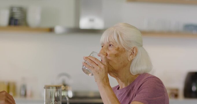 Senior woman drinking from glass at kitchen counter after hand offering white bottle with red cap
