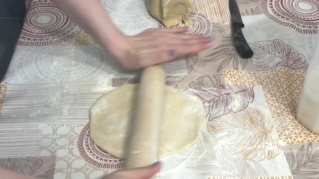 A woman flips and rolls out raw dough with a rolling pin on a table, home kitchen setting. Homemade pastry preparation, traditional baking, culinary process.