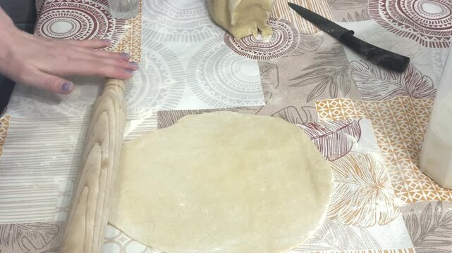 A woman flattens raw dough with a wooden rolling pin on a patterned tablecloth, home kitchen setting. Homemade pastry preparation, traditional baking, culinary process.