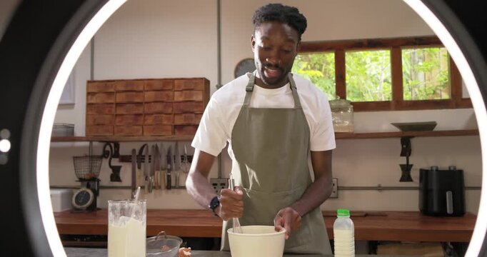 Ring light turning on, African man in olive apron whisking batter at counter, demonstrating recipe