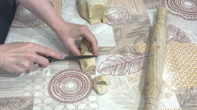 A woman carefully slices a roll of dough into small equal portions using a knife on a table, kitchen environment. Small batch baking, pastry making, cozy home cooking.