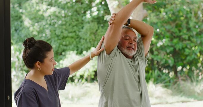Asian PT guiding senior African American man lifting arm overhead in yard for shoulder care, watch
