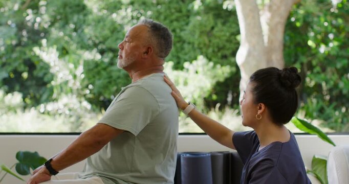 Senior man in rehab-clinic rotating neck and torso as Asian clinician placing hands by foam rollers