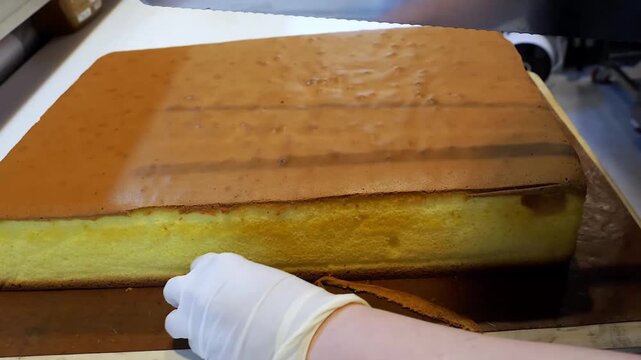 Close-up shot of a person wearing white gloves using a large knife to cut a slice from a giant, golden-brown Japanese or Taiwanese-style Castella sponge cake on a wooden board.
