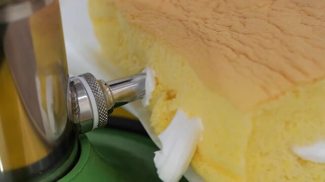 A close-up shot captures a metal piping tip carefully inserting fresh white cream into a slice of a golden-yellow, bouncy Taiwanese Castella cake. The focus is on the soft, airy texture of the sponge 