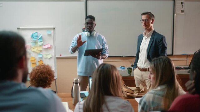 African male student presenting project with tablet during university class while professor facilitates discussion and engages other students. Teamwork, brainstorming and collaborative learning.
