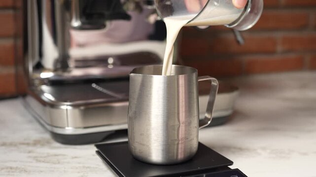 Barista pouring milk into metal pitcher on digital scale. Measuring precise amount of milk for coffee preparation in cafe