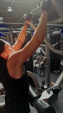 Vertical shot of a young man exercising on lat pulldown machine in a bright gym. Fitness, strength training and healthy lifestyle concept with natural daylight. High quality 4k footage