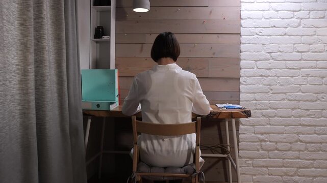 Woman seated with back to camera, focused on laptop at live edge wooden desk in modern home office with exposed brick wall, cozy, productive remote work setup