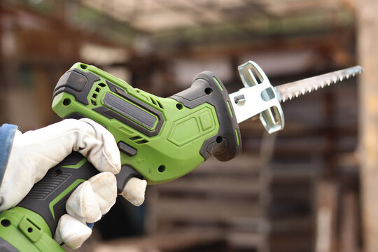 A craftsman in a protective glove holds a compact green cordless reciprocating saw. The tool is designed for fast and efficient sawing of wood, metal, plastic and plywood. Power tool.