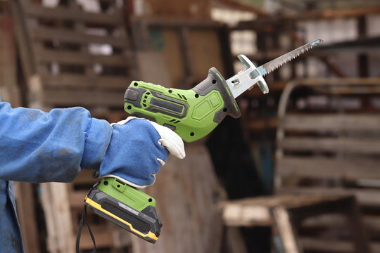 A craftsman in a protective glove holds a compact green cordless reciprocating saw. The tool is designed for fast and efficient sawing of wood, metal, plastic and plywood. Power tool.