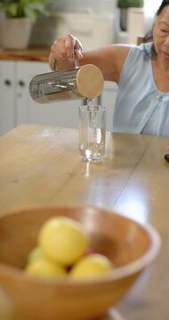 Vertical video: Senior Asian woman tilting pitcher pouring into glass drinking at table, copy space