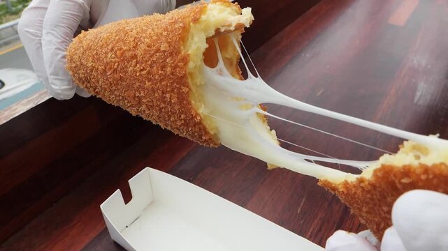 A person wearing white gloves pulls apart a freshly fried, crispy Korean corn dog, revealing the satisfyingly stretchy, melted mozzarella cheese filling. This popular and trendy street food snack is c