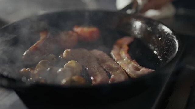 Smoke rises from a hot pan of sizzling bacon. Tongs turn crispy strips over sizzling mushrooms. The sizzle hints at savory breakfast or brunch. Use for food ads, cooking tutorials