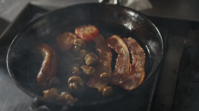 Sizzling bacon and mushrooms sizzle in a black skillet. Steam curls upward as the food cooks. Tongs gently flip the sausages in the pan. Close-up of a hearty breakfast meal. The sizzle conveys warmth
