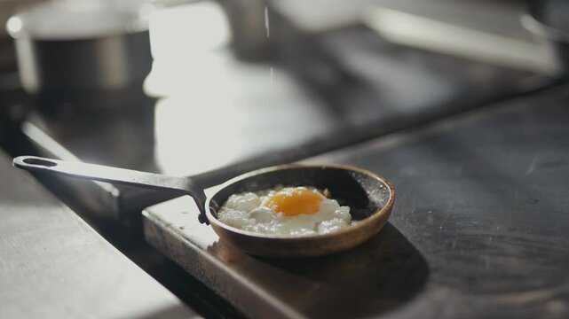 Soft light glows over a tiny pan. Fried egg rests gently in the skillet. Wooden board holds the pan. Warm, rustic kitchen setting. Ideal for food or lifestyle content. No people or animals visible