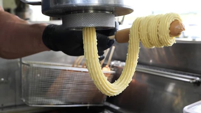 Close-up shot of a person wearing a black glove using a churro machine to wrap dough around a hot dog on a stick, preparing a unique sweet and savory snack for deep frying.