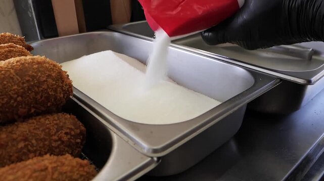 Close-up footage of a person wearing black gloves pouring a white, granular substance, likely sugar, from a red and white bag into a stainless steel preparation tray. In the foreground, several golden
