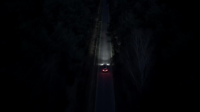 Aerial night shot of a car driving through dark forest with headlights on as drone follows vehicle along straight road