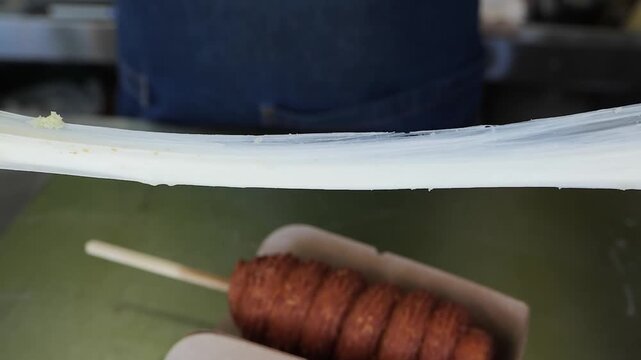 Close-up of a person wearing black gloves pulling apart a freshly fried Korean cheese corn dog on a stick, highlighting the long, melted mozzarella cheese pull. Another plain corn dog sits in a paper 
