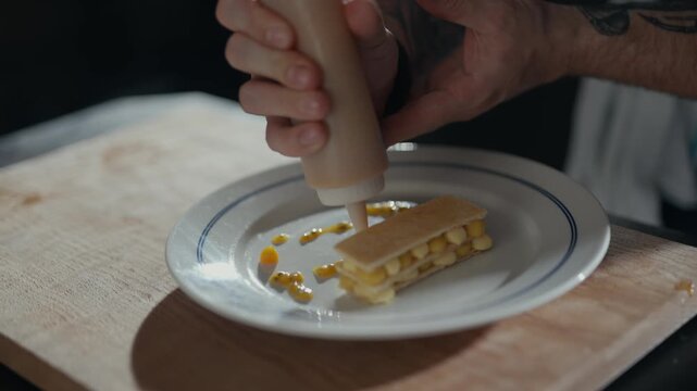 Hands squeeze sauce over a plated dish. Fingers steady as a condiment drizzles from a squeeze bottle. Plate holds a neatly arranged, square-shaped food item. Background shows a wooden cutting board