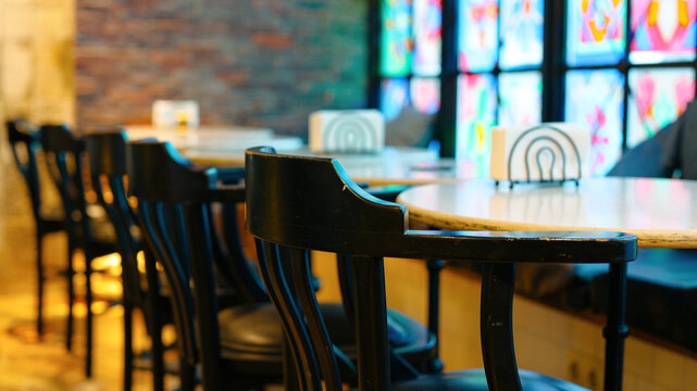 Empty chairs lining wooden counter tables inside a modern cafe or bar with a blurred background of colorful stained glass windows, creating a cozy and inviting atmosphere
