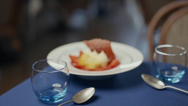 Dinner plate with dessert, blue tablecloth, and glasses. Spoon and fork rest beside the dish. Soft focus creates an intimate, cozy restaurant scene. Ideal for food, lifestyle
