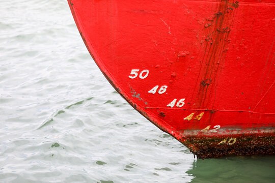Red bow showing draft marks and waterline on a large vessel, indicating its displacement and cargo capacity, resting calmly in the port's water