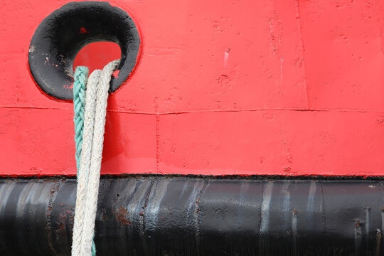Vessel hull close up showing a section of the red painted metal hull, featuring a black hawsehole passing two thick ropes, one green and one white, securely mooring the boat