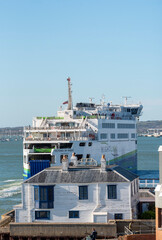 Fototapeta premium A large passenger and vehicle ferry, the Victoria of Wight, navigates the water near a quaint white house with blue window frames in Portsmouth Harbour UK