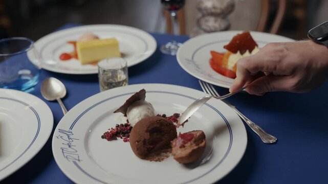 Table set with dessert plates and wine glasses. Hand uses spoon to scoop into a chocolate dessert. Blue tablecloth and white porcelain plates. Scene suggests intimate, quiet dining experience