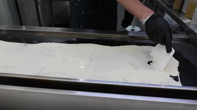 close-up shot of a professional chef wearing black gloves using a flat spatula tool to spread or level a smooth, white substance, possibly dough, batter, or cream, across a clean metallic kitchen work