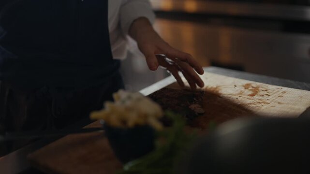 Chef's hands slice meat on wooden board. Fingers show skillful, precise movements. Dark background highlights kitchen activity. Professional cooking scene, close-up view. Ideal for food, culinary