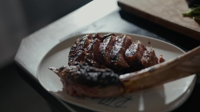 Hand slices meat with care. Sear marks glow under soft light. Bone-in steak rests on a plate. Food is prepared for a meal. Warm atmosphere suggests comfort. Ideal for dining or cooking scenes