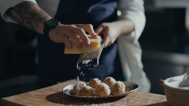Chefs hands grate cheese over golden fried balls. Warm kitchen light highlights the motion. Fresh cheese dusts the dish with care. Texture of food creates an inviting look