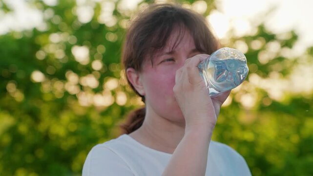 Tired woman drinks refreshing water from bottle, walk, park, nature. Woman feeling unwell drinks pure mineral water outside, summer heat in park. Exercise outdoors, quench your thirst with cool water