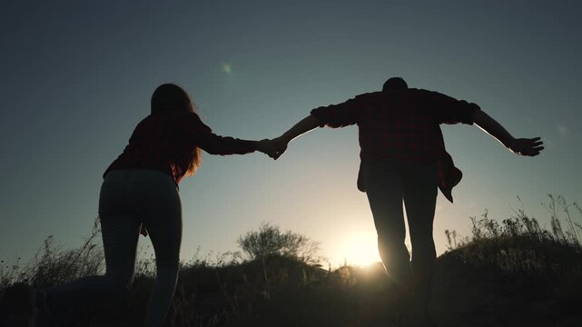 Couple holding hands running on hill at sunset. Silhouette of man and woman run together outdoors. Partners hold hands during sunset run on hillside. Couple enjoys outdoor adventure at dusk.
