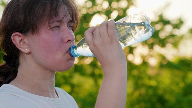 Tired woman drinks refreshing water from bottle, walk, park, nature. Woman feeling unwell drinks pure mineral water outside, summer heat in park. Exercise outdoors, quench your thirst with cool water