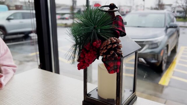 A decorative holiday lantern with pine, berries, and pinecone on a table by a window.