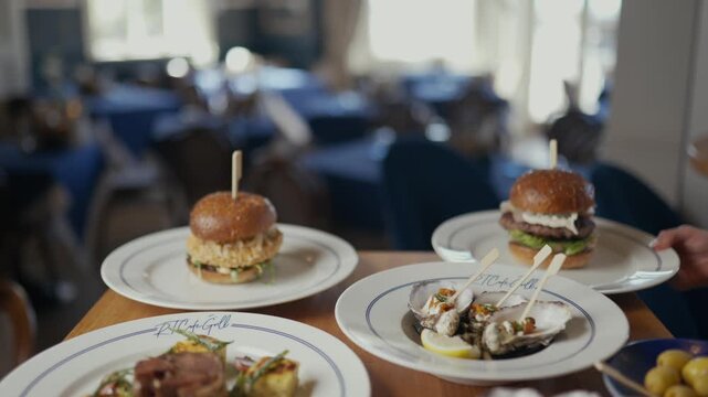 Wooden table holds plates of food. Small burgers with toothpicks sit ready to eat. Oysters and garnishes complete the meal. Soft focus shows empty dining area behind. No people appear in the scene