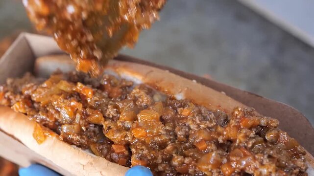 A close-up, appetizing shot of a person wearing a blue protective glove, holding a fresh, hot chili dog. The hot dog is nestled in a soft bun and generously topped with a rich, steaming meat sauce (ch
