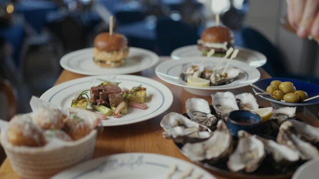 Hand pours condiment over oyster plate. Oysters rest in open shells, served on metal tray. Small burgers and side dishes surround the seafood. Dinner scene evokes freshness, luxury, and casual dining