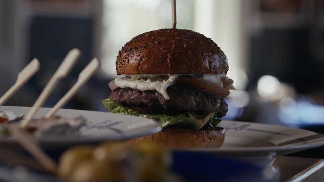 Delicious burger with toothpick sits on plate. Garnished with greens and melted cheese. Background shows skewers and small potatoes. Food looks fresh and savory